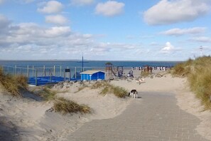 Ein Strandweg führt zu einer blauen Schutzhütte, einem Rettungsschwimmturm und dem Meer im Hintergrund.