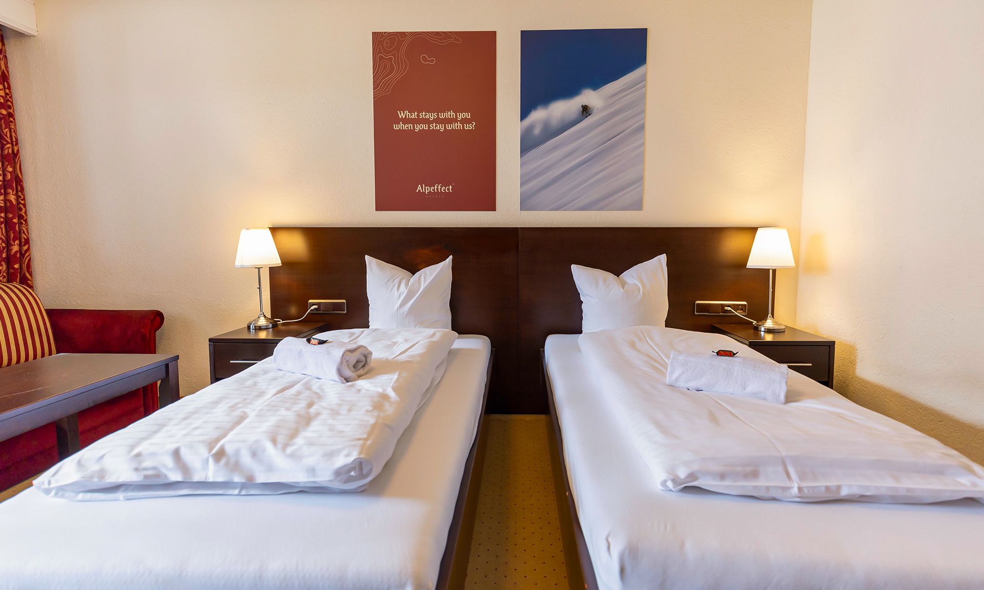 Een hotelkamer met twee bedden, elk voorzien van een kussen en een opgevouwen handdoek, een nachtkastje met een lamp, en een muur met twee posters.