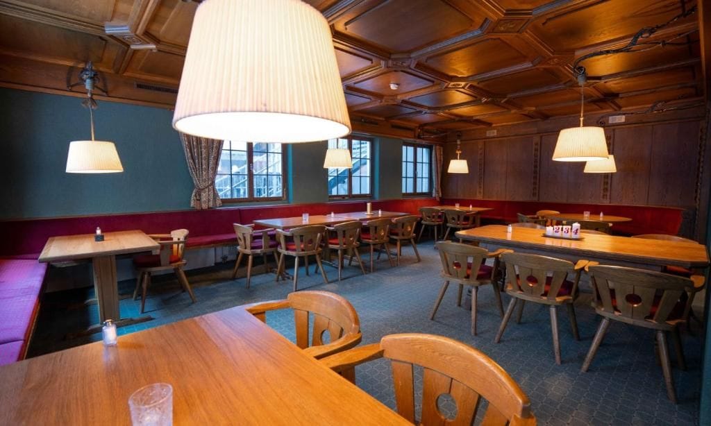 Un restaurant avec des tables et des chaises en bois, de grandes lampes suspendues et une vue sur l’extérieur par les fenêtres.