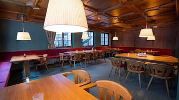 Un restaurant avec des tables et des chaises en bois, de grandes lampes suspendues et une vue sur l’extérieur par les fenêtres.