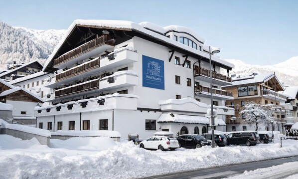 A multi-story hotel with a prominent sign for Hotel Rosanna, surrounded by snow-covered buildings and a mountainous backdrop.