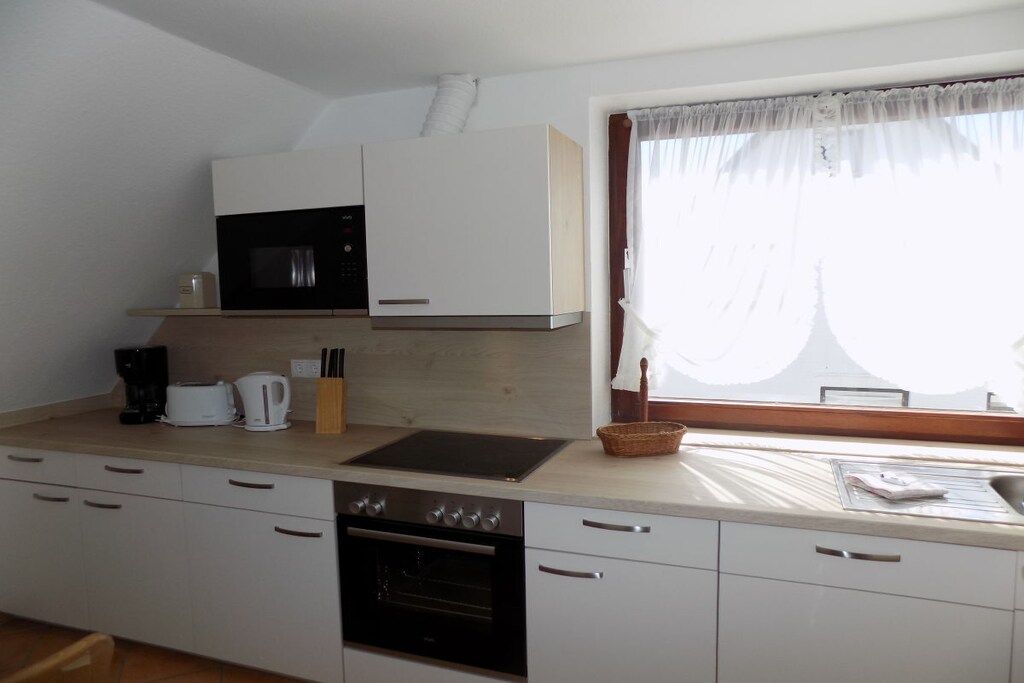Une cuisine avec des meubles blancs, un four à micro-ondes, une cuisinière et une fenêtre ornée de rideaux légers.