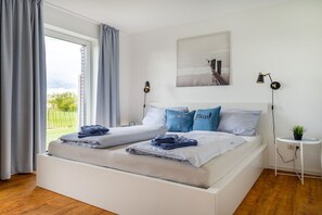 A double bed with a view of a garden through a window, a bedside table with a lamp, and a wall-mounted artwork. - Meerhuus Carolinensiel House 2 (Carolinensiel)