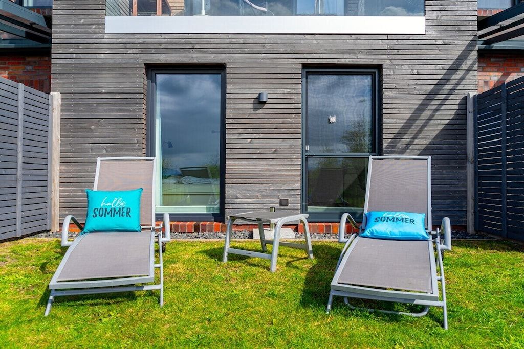 Dos sillones con almohadas que dicen 'hello SOMMER' frente a un edificio moderno con ventanas amplias.