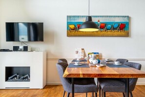 Un saló modern amb televisió de pantalla plana, una llar de foc, una zona de menjador amb taula de fusta i cadires grises, i un quadre amb temàtica de platja sobre la taula.