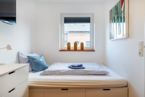 Ein Schlafzimmer mit einem Einzelbett, einem Fenster mit Jalousien, einem an der Wand befestigten Fernseher und einem kleinen Nachttisch.