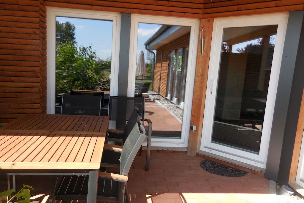 Eine Holzdielen-Terrasse mit Tisch und Stühlen, eine Schiebeglastür und Blick auf einen Garten.
