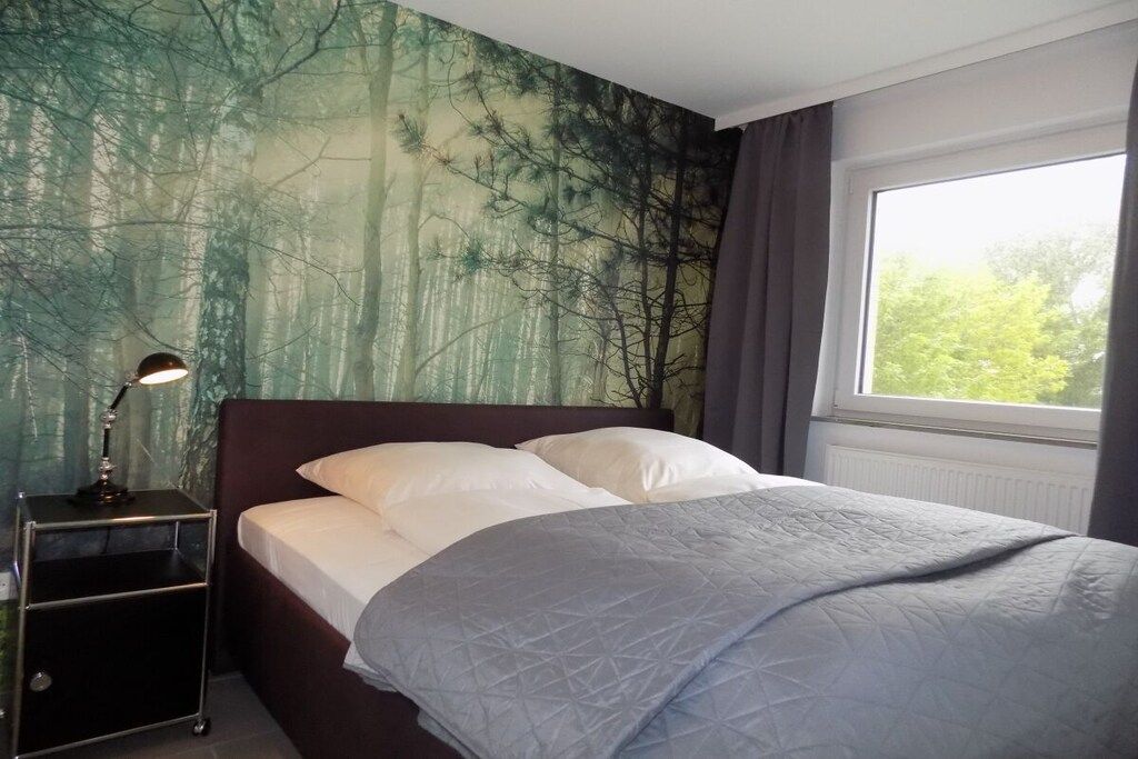 Une chambre à coucher avec un lit, une table de chevet, une fenêtre et une fresque représentant une forêt sur le mur.