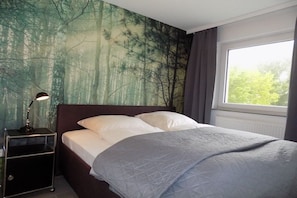 A bedroom with a bed, a nightstand, a window, and a forest mural on the wall. - Theodor-Heuss-Str. Holiday Apartment 11 (Westerland)