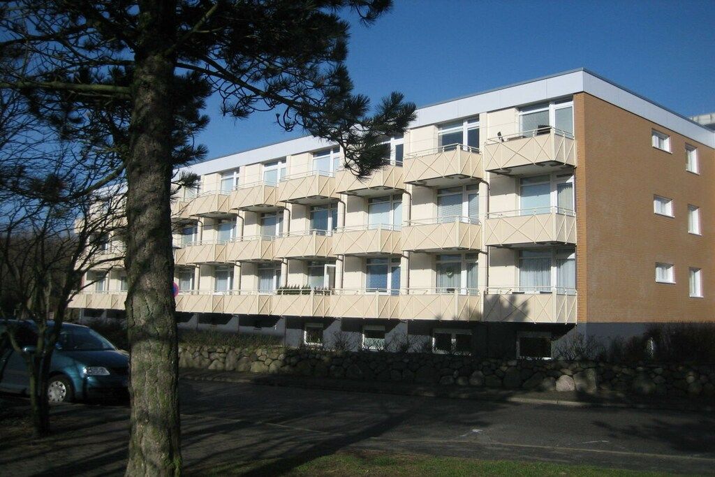 Un edificio a più piani con balconi, un muro di pietra di fronte e un'auto parcheggiata.