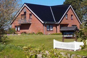 Una casa a due piani in mattoni con tetto inclinato, un balcone al piano superiore e un piccolo giardino recintato da una staccionata bianca.