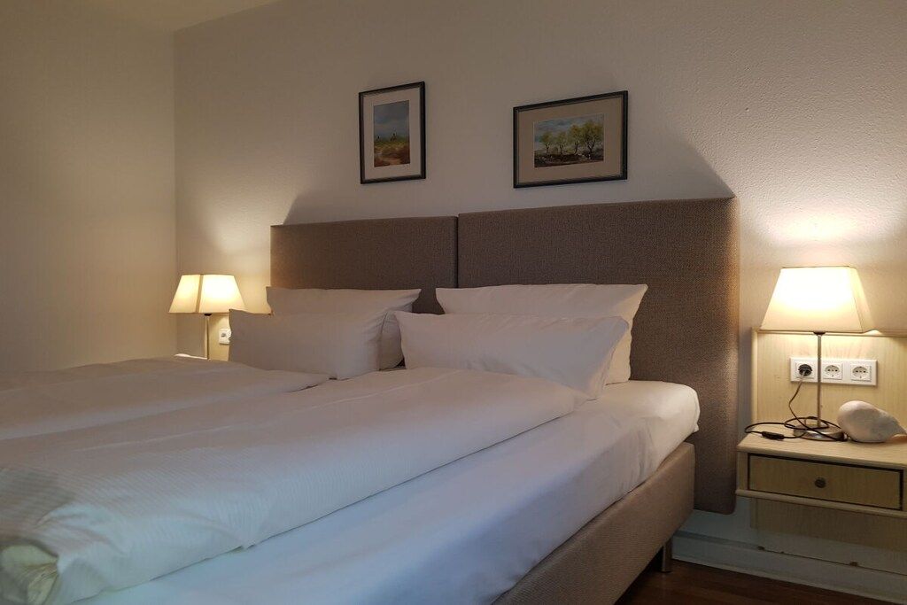 Ein ordentlich bezogenes Bett mit weißen Leinen, einem Kopfteil, Nachttischlampen und zwei gerahmten Bildern an der Wand.