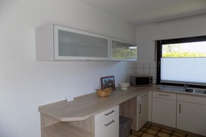 A compact kitchen with a microwave, a sink, and a window with blinds. - Ground floor holiday apartment Zur Eiche 19 (Tinnum \/ Sylt)