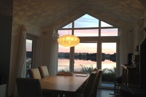 A dining area with a table and chairs, a hanging light fixture, and large windows overlooking a body of water. - Marina Hülsen - The Sonnenuferhaus (Kosel)