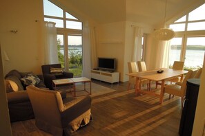 A living room with a sofa, a coffee table, a TV, a dining table with chairs, and large windows overlooking a body of water. - Marina Hülsen - The Sonnenuferhaus (Kosel)