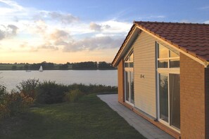 A house with a sloped roof, large windows, and a view of a lake with a sailboat. - Marina Hülsen - The Sonnenuferhaus (Kosel)