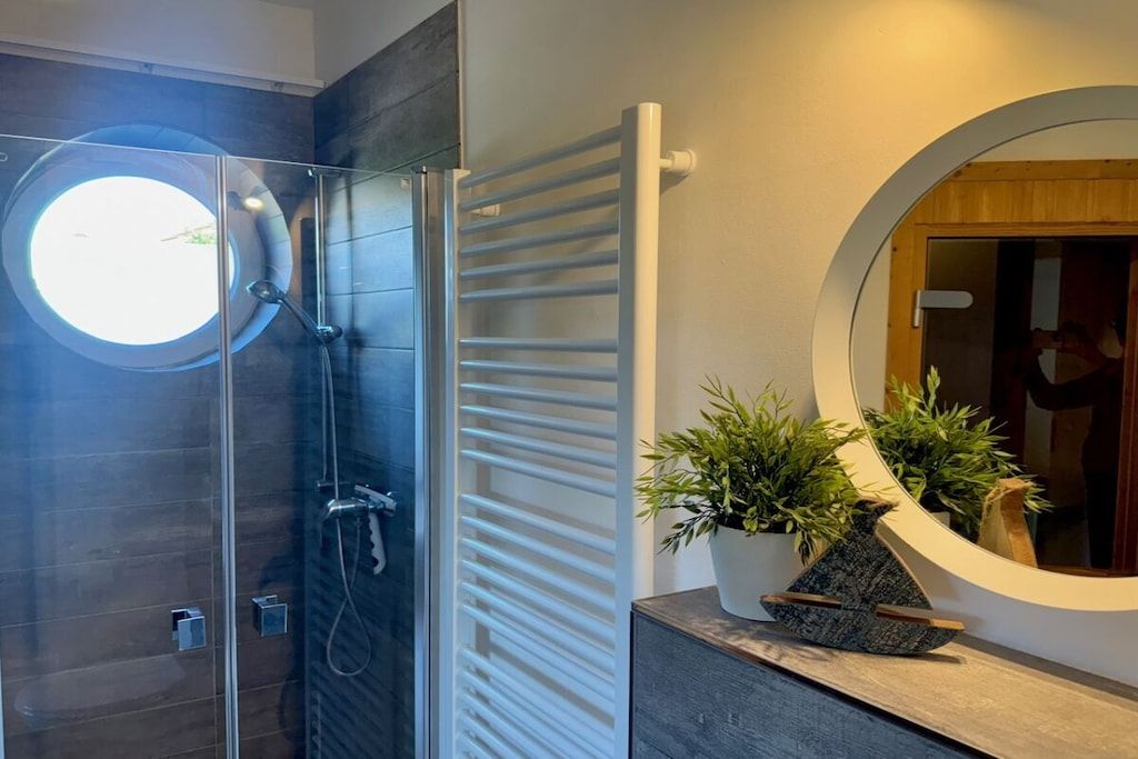 Un baño moderno con ducha envuelta en vidrio, toallero blanco, espejo redondo y planta en un mueble de tocador de madera.