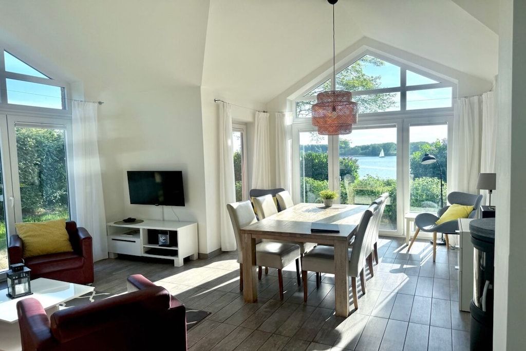 Ein modernes Wohnzimmer mit großem Fernseher, Esstisch und Stühlen, großen Fenstern mit Vorhängen und Blick ins Freie.