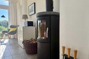 Une pièce avec un poêle à bois, une chaise avec un coussin jaune, un panier en osier et un bureau avec une lampe.