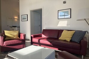Un salon avec un canapé rouge, une table basse blanche et un fauteuil rouge avec un coussin jaune. Une porte donne accès à une autre pièce, et un tableau encadré est accroché au mur.