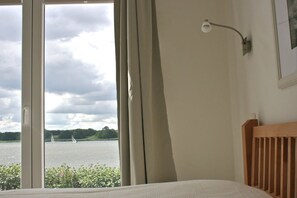 A bedroom with a large window, a wooden headboard, and a wall-mounted lamp. - Marina Hülsen - The Reed Bank House (Kosel)