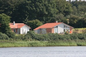 Two houses with red roofs, white walls, and large windows, situated by a body of water with dense greenery in the background. - Marina Hülsen - The Reed Bank House (Kosel)