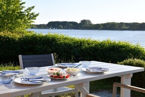 An outdoor dining table set with plates, bowls, and cutlery, overlooking a body of water. - Marina Hülsen - The Swan Embankment House (Kosel)