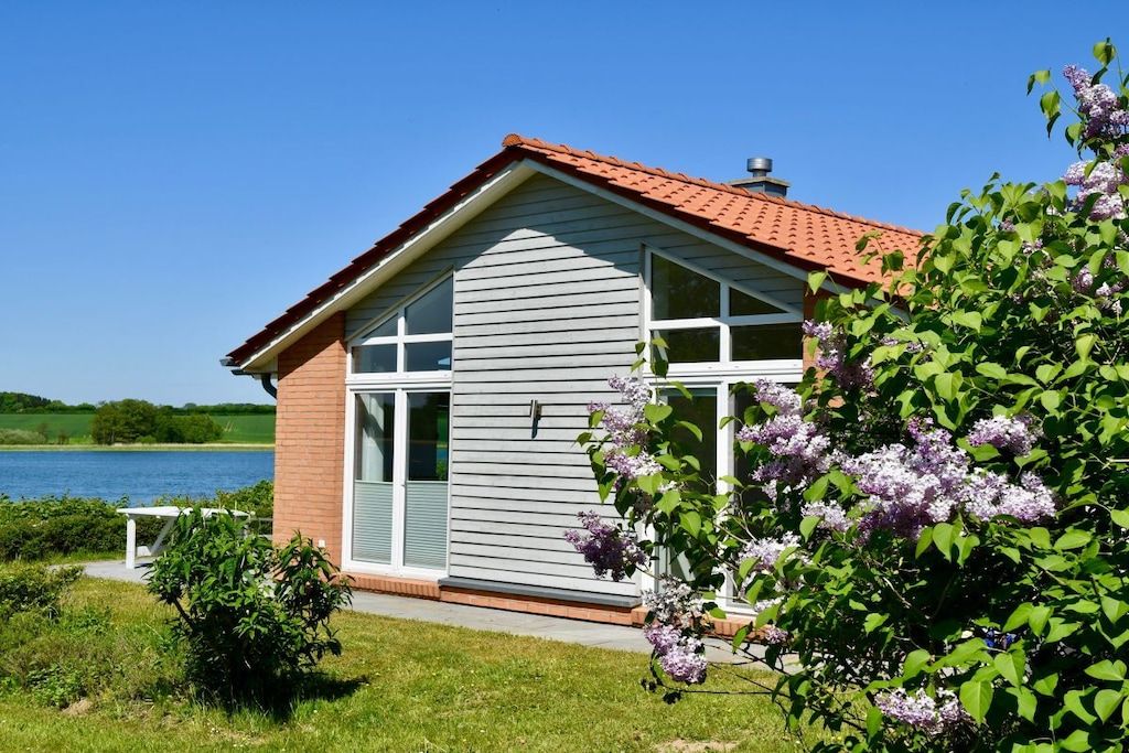 Una casa con techo rojo, ventanas grandes y vista a un lago.