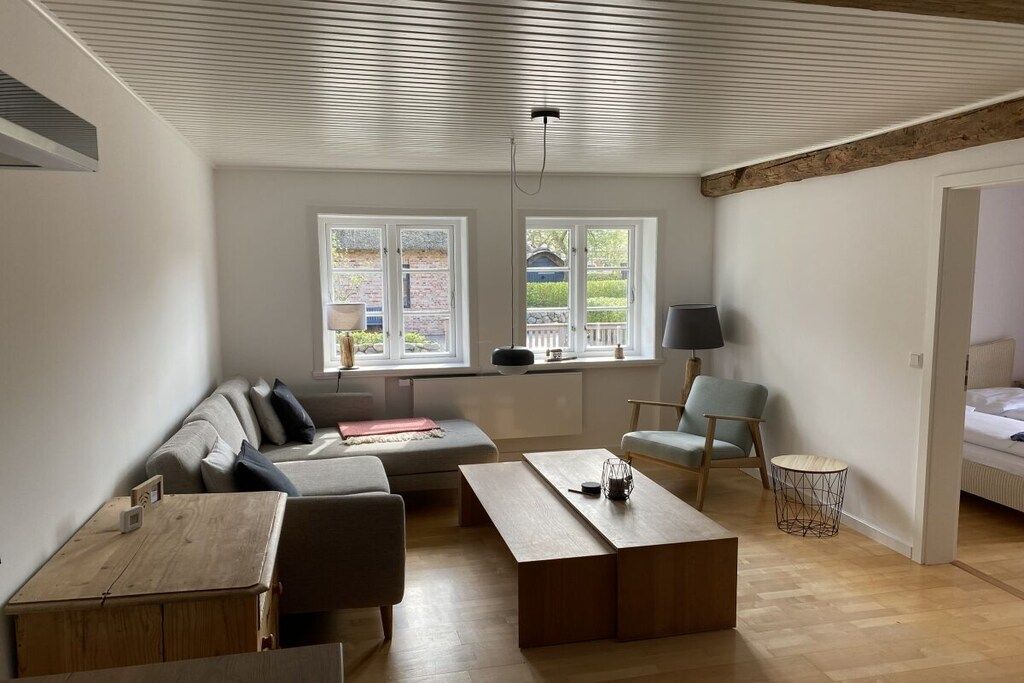 A modern living room with a sofa, a coffee table, and a chair. There are two windows with a view of the outside.