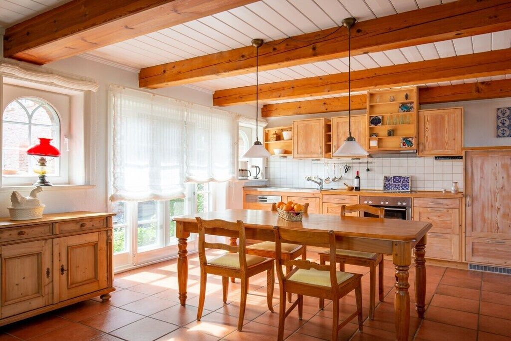 Een keuken met houten kastjes, een eettafel en stoelen. Er is een raam met gordijnen en een rode lamp op een bijzettafeltje.