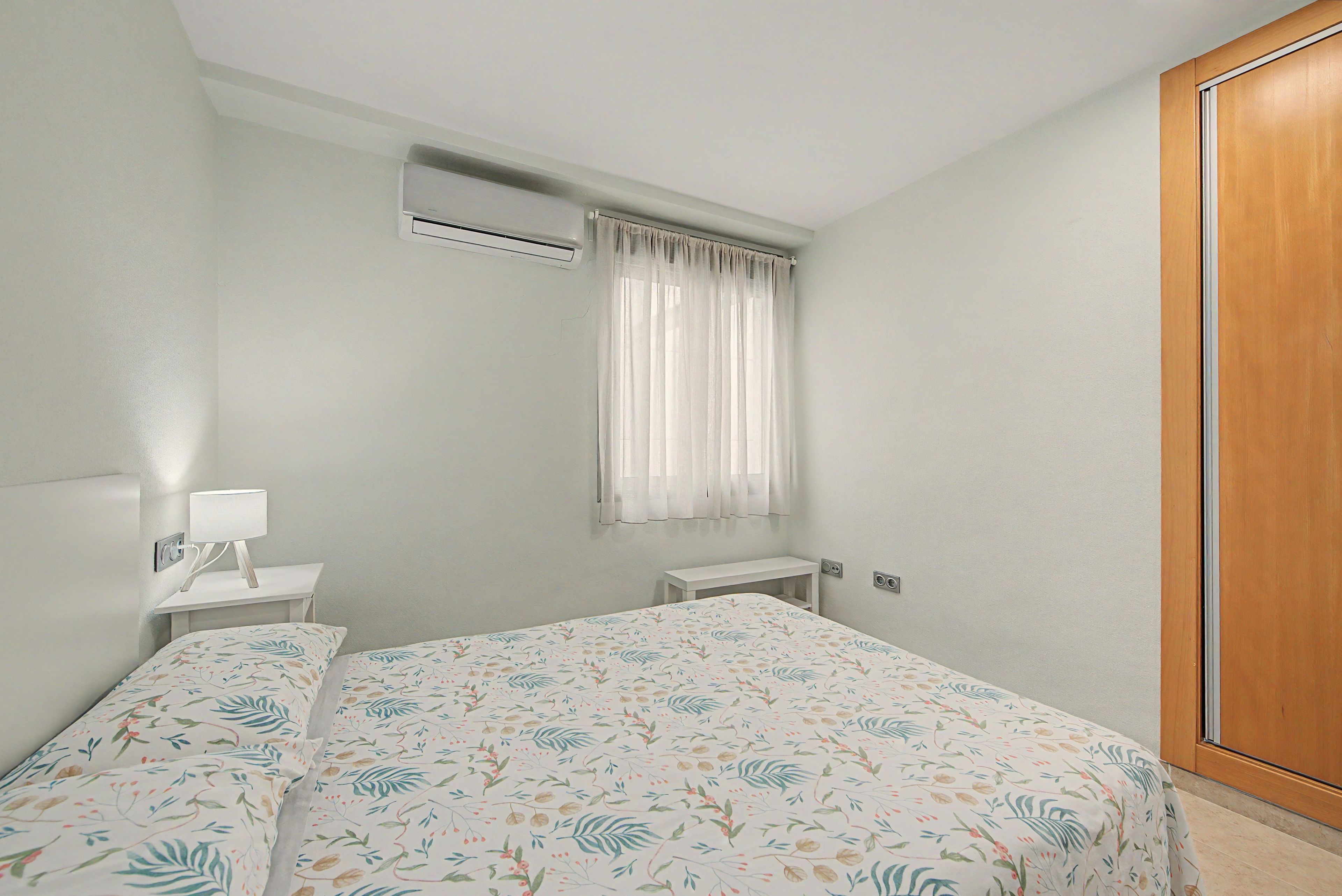 Una camera da letto con un letto, un comodino con una lampada, un condizionatore d'aria, una finestra con tende e un armadio in legno.