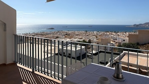Uma varanda com mesa e cadeiras, com vista para um horizonte costeiro com edifícios e o mar.
