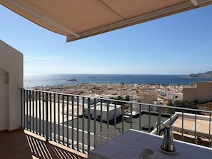 En balkon med et bord og stole, udsigt over et kystlandskab med bygninger og havet.