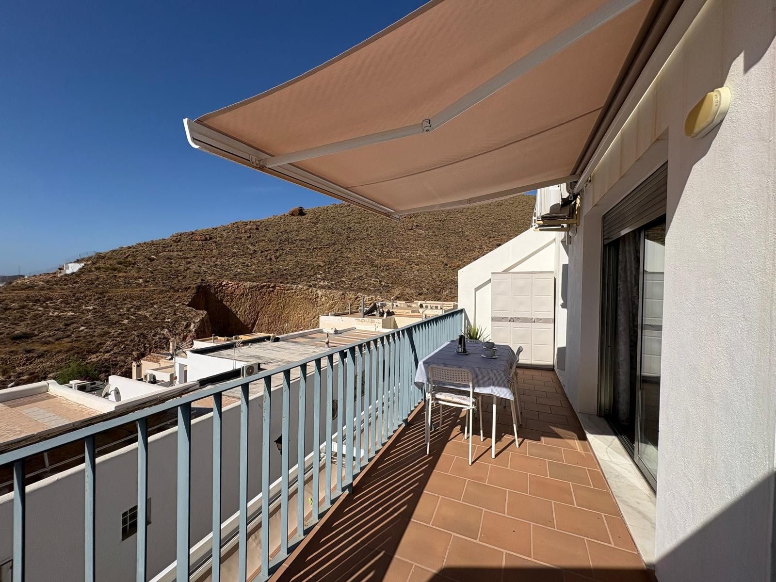 A balcony with a table and chairs, a white building, and a view of a hilly landscape.