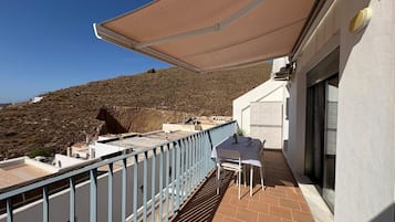 A balcony with a table and chairs, a white building, and a view of a hilly landscape.