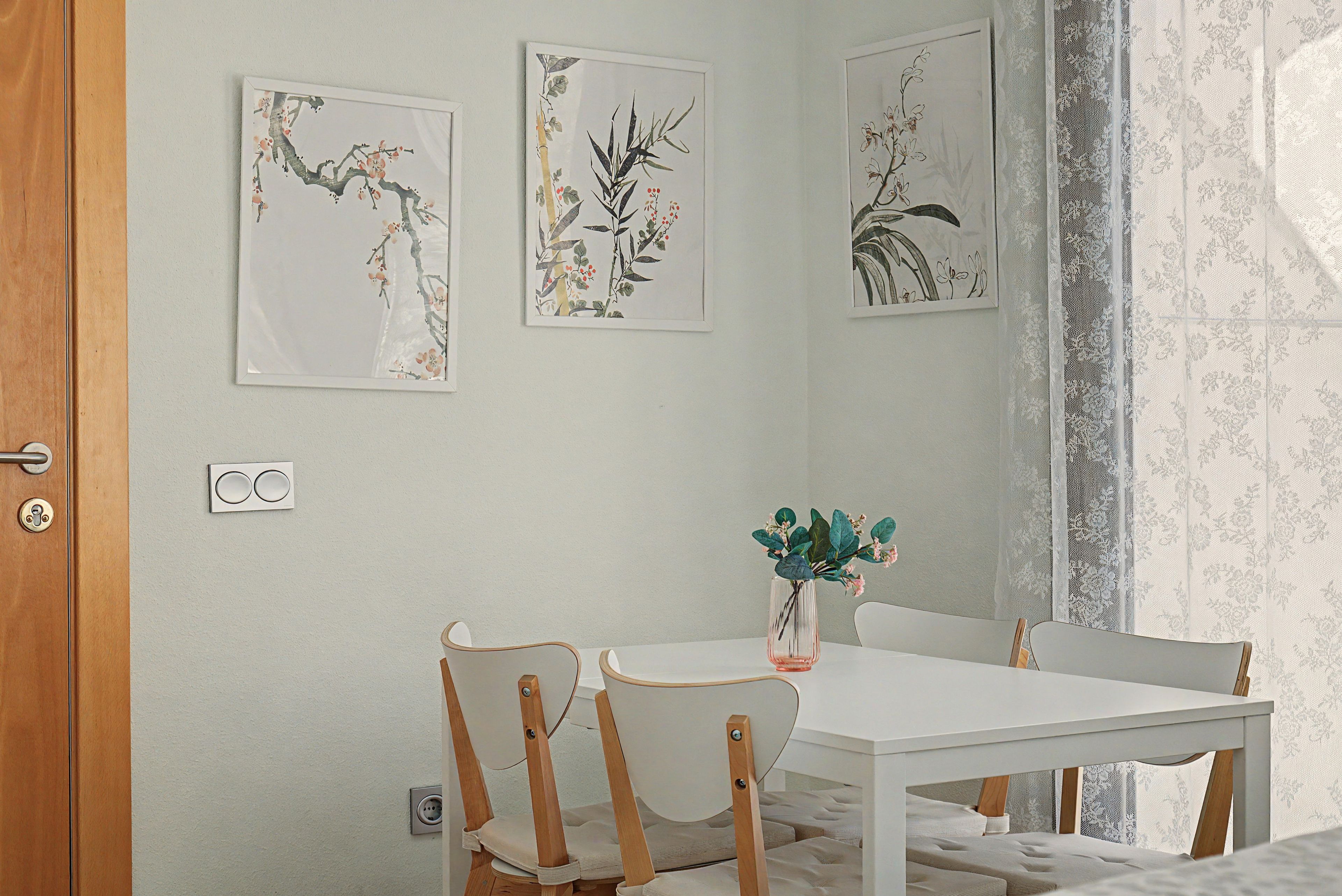 Zona pranzo con tavolo e sedie bianche, un vaso con fiori e tre stampe botaniche incorniciate al muro.