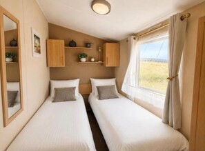 A double room with two beds, a mirror, a shelf with decorative items, and a window with curtains. - Seaside Escape at St Osyth (Clacton-on-Sea)