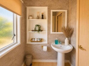 A bathroom with a window, a sink, a mirror, and shelves with toiletries. - Seaside Escape at St Osyth (Clacton-on-Sea)