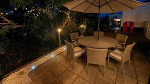 Un patio exterior con una mesa redonda y sillas, rodeado por un muro bajo y luces colgantes.