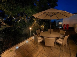 An outdoor patio with a round table and chairs, surrounded by a low wall and string lights. - Seaside Escape at St Osyth (Clacton-on-Sea)