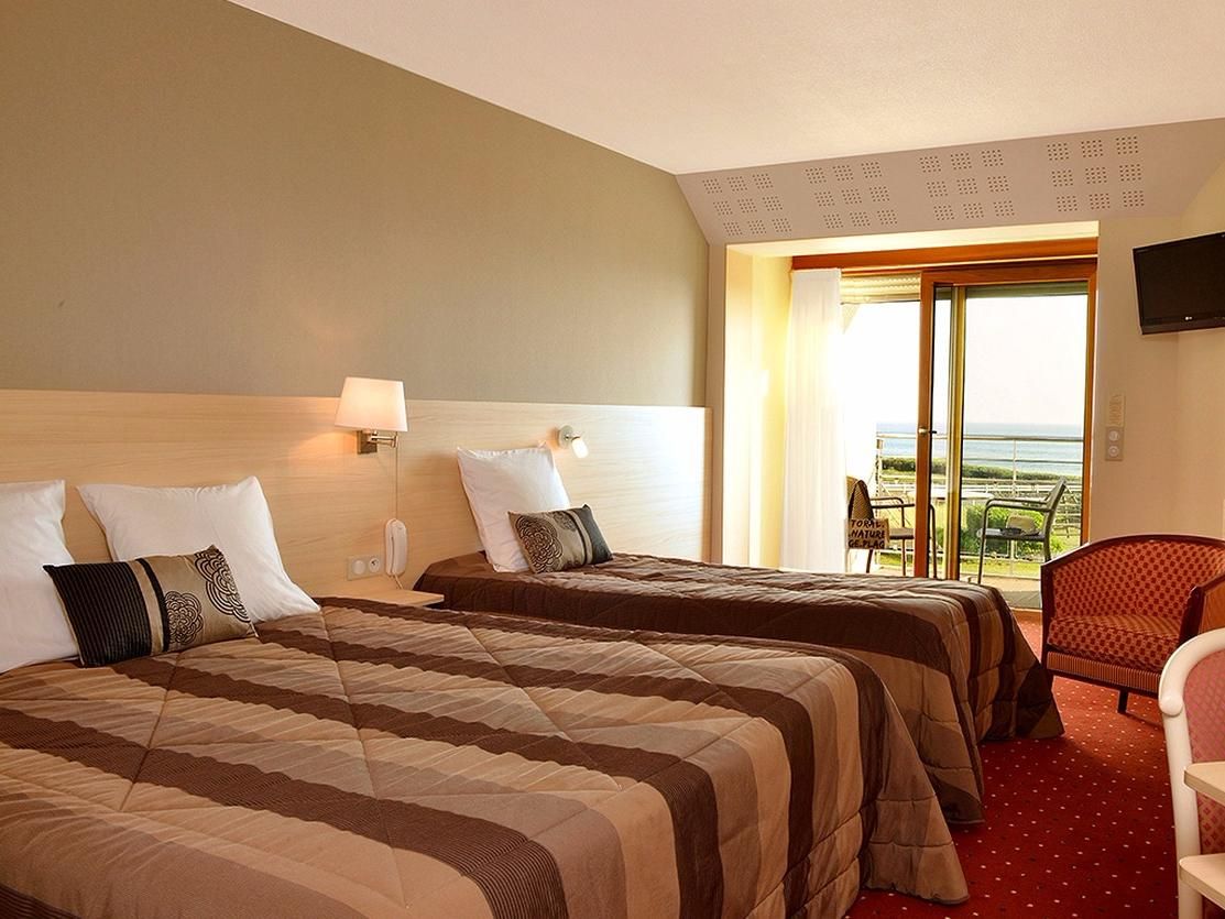 A hotel room with two beds, a red carpet, a TV on the wall, and a view of the sea.