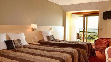 A hotel room with two beds, a red carpet, a TV on the wall, and a view of the sea.