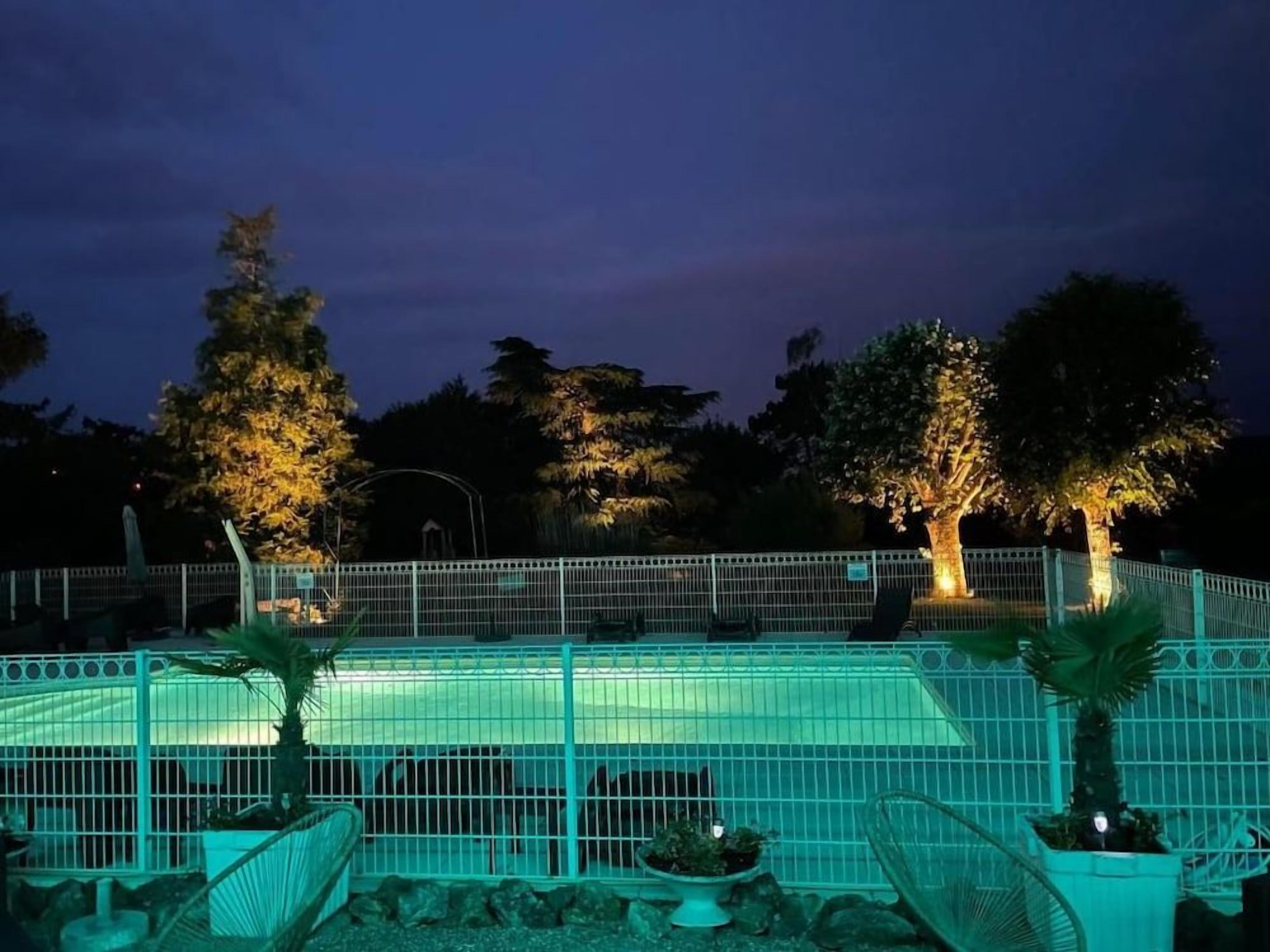 Un espace extérieur clos, doté d’une piscine, entouré d’arbres et éclairé par un système d’éclairage artificiel.