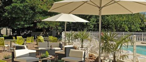 An outdoor seating area with wicker furniture and umbrellas, a pool in the background, and a white fence surrounding the space.
