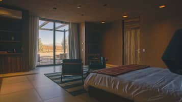 A modern bedroom with a large bed, a desk, and a chair. There is a sliding glass door leading to an outdoor area.