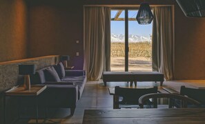 Una habitación con sofá, banco, mesa y vista a las montañas a través de un ventanal grande.