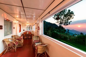 A corridor with a dining area featuring wicker chairs and a chessboard, leading to a window with a view of a mountainous landscape.