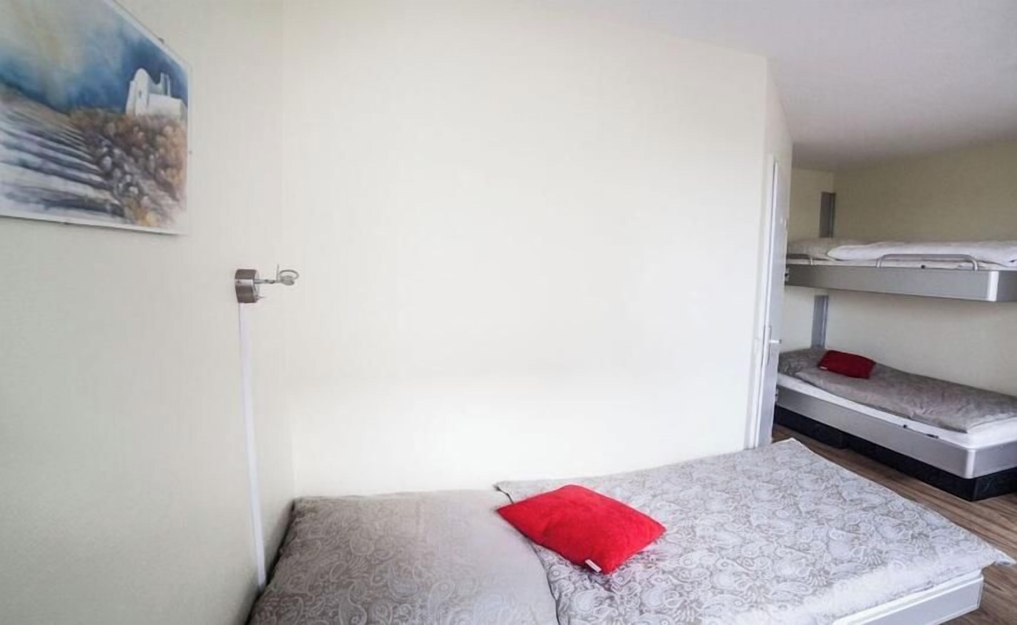 Ein Einzelbett mit einem roten Kopfkissen, ein Etagenbett im Hintergrund und ein Gemälde an der Wand.