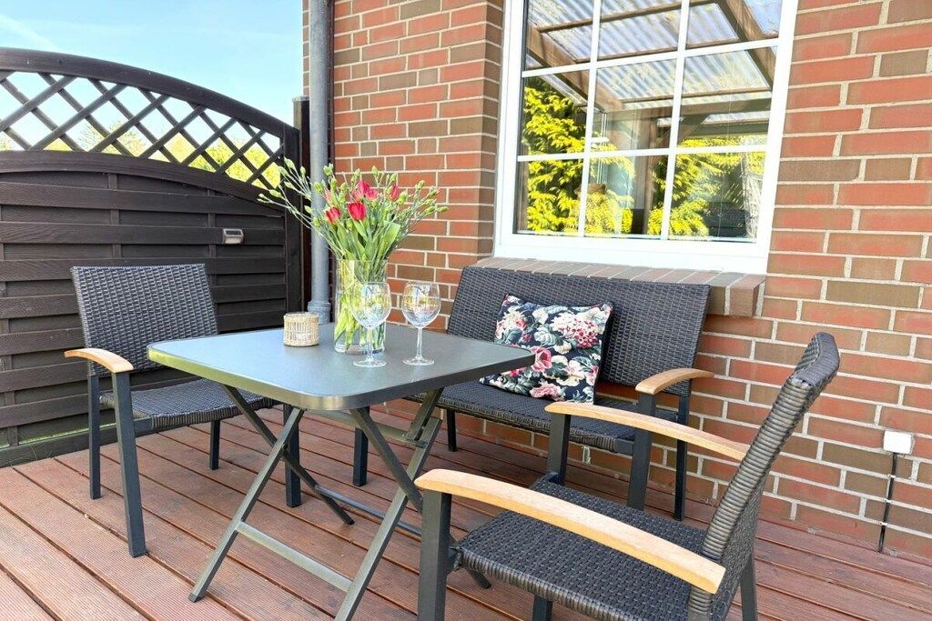 An outdoor patio with a table and chairs, a vase with flowers, and a window with a view of trees.