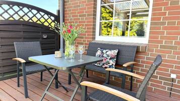 An outdoor patio with a table and chairs, a vase with flowers, and a window with a view of trees.